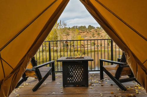 Cozy tent interior with two chairs and a fire pit, overlooking a scenic view of trees and water.